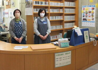 木製カウンター越しに図書館の職員が案内用デスクに立ち、背後の書架が整然と並ぶ様子の写真
