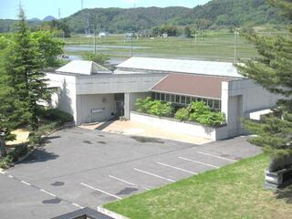 山と田園の風景を背景に建つ、平屋建てで灰色の壁と一部茶色の屋根を持つ現代的な建物の前に広い駐車場がある図書館の外観写真