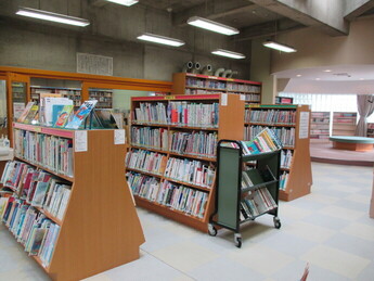 明るい照明の屋内空間で、高さの異なる木製書架に、たくさんの児童書や絵本が表紙を見せて並べられており、奥には円形のステップ状の読書スペースが見える様子の写真