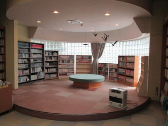 円形の小さなステージ空間に丸いベンチと周囲を囲む書架、床置きの暖房器具が配置された落ち着いた読書エリアの写真