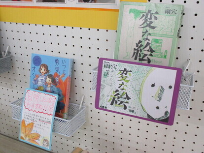 白い有孔ボードに設置されたラックに、「変な絵」と女の子2人のイラストが表紙の図書が手書きの紹介ポップと共に並べられている写真