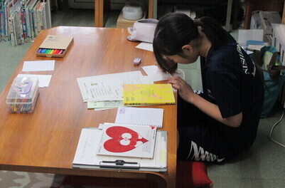 資料やカラーペンが置かれたテーブルに向かってポップを作っている女子生徒の写真