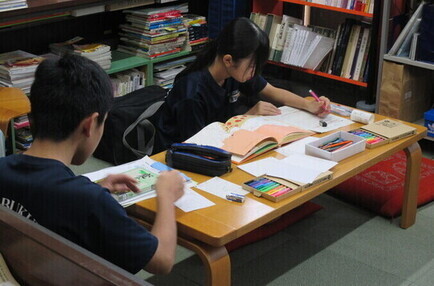 本やカラーペンが置かれたテーブルに向かって、男女二人の生徒がポップを作っている写真