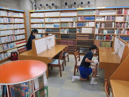 壁一面に並んだ本棚に囲まれた図書室で、紺色の服を着た2人の女子生徒が机や棚の清掃に取り組んでいる様子の写真