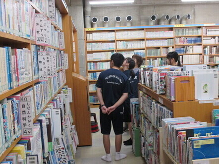 たくさんの図書が並ぶ本棚を見上げながら、司書が数名の生徒に本の並ぶ順番を説明している写真