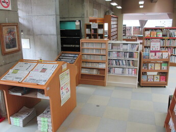 図書館の新聞コーナーの手前に新聞を広げて閲覧できる傾斜した木製の台が置かれ、その後ろにDVDなどが並んだ木製の書棚が並んでいる写真