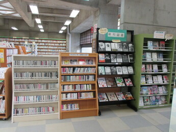 コンクリート打ちっぱなしの壁と天井の図書館の一角で、左からCDが並んだ白い棚、木製棚、新刊図書や雑誌が並ぶ黒と緑の陳列棚が並んでいる様子の写真