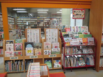ガラスの壁の前に、絵本や読み聞かせ本などが表紙を見せて陳列された木製と金属製の展示棚が並んでいる写真