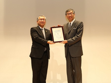 スーツ姿の二人の男性がステージ上で並んで立ち、額に入った登録証を両側から持っている写真