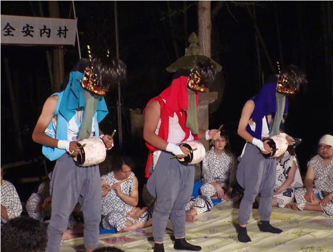 夜の境内で色鮮やかな装束と仮面を着けた三人が太鼓を打ち鳴らして舞う様子の写真
