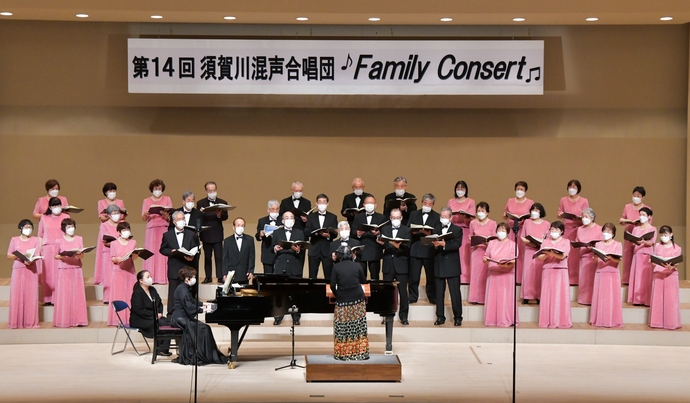 「第14回 須賀川混声合唱団 Family Consert」という看板が掲げられたステージ上で、ピンクのドレスを着た女性陣と黒のタキシード姿の男性陣が並び、ピアノ伴奏に合わせて合唱を披露している華やかなコンサート風景の写真
