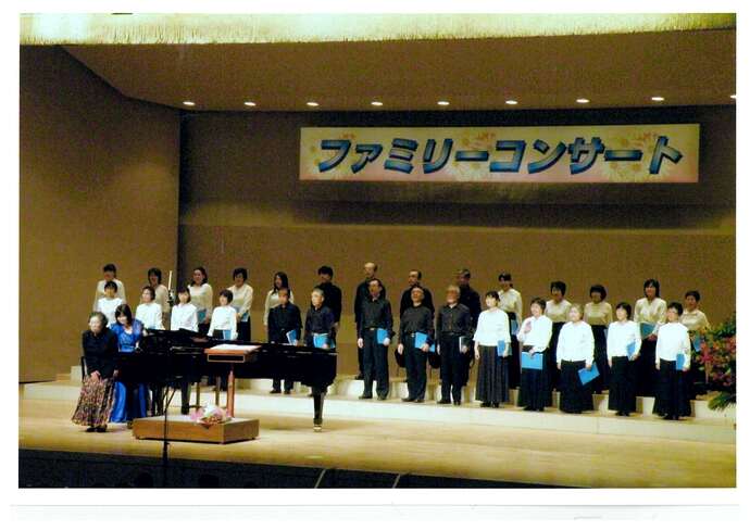 「ファミリーコンサート」という看板が掲げられた舞台の上で、白や黒の衣装に身を包んだ合唱団が整列し、ピアノの横に立った2人の女性と共に客席を向いている写真