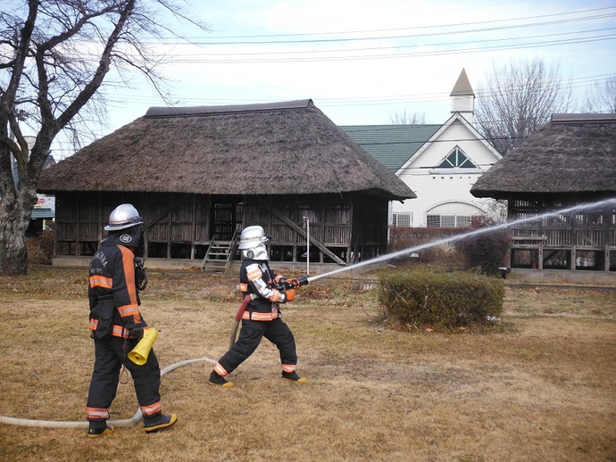 ヘルメットと防火衣を着用した2人の消防団員が、茅葺き屋根の歴史的な建物の前でホースを構え、訓練のために勢いよく放水を行っている、冬の枯草が広がる広場での活動風景を捉えた写真