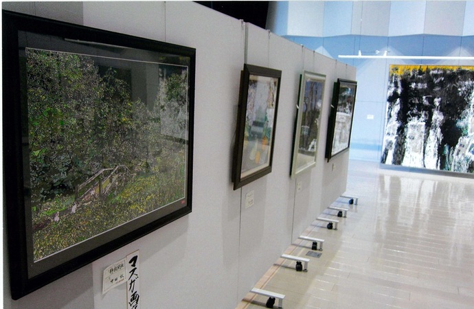 明るく清潔感のある展示会場にて、白いパーテーションに等間隔で並べられた風景画などの額装作品と、その奥の壁面に飾られた大きな抽象画を斜めの角度から見渡すように撮影した写真