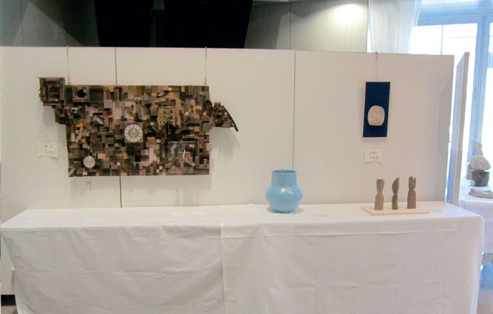 白い展示用パーテーションに掛けられた、木片を組み合わせたような複雑な造形の時計付きの作品や、手前の白い布が敷かれた展示台に置かれた水色の花瓶や小さな立体作品を正面から撮影した写真