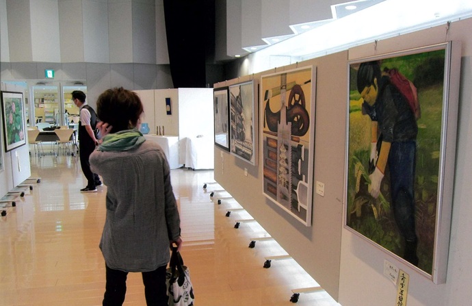 複数の絵画作品が並ぶ白いパーテーションの間の通路を男女の来場者が鑑賞している、展覧会の様子の写真