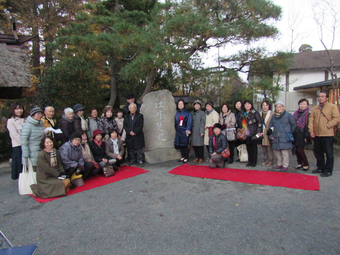 建物の前の木々を背景に、中央にある大きな石碑を囲んで、20名以上の男女が笑顔で並んで記念撮影をしている写真
