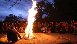 夜の暗闇の中で勢いよく燃え上がる巨大な焚き火を、大勢の人々が静かに囲んで見守り、火の粉が舞い上がる幻想的な光景と、地面を照らす暖かな光が印象的な屋外での行事の様子を捉えた写真