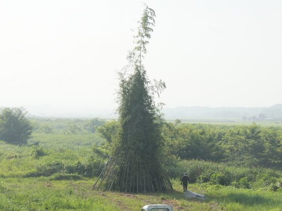 青竹を幾重にも組み合わせて円錐状に高く積み上げた大きなやぐらの足元で作業者が器具を手に準備を進め、その壮大な竹構造の高さと形状の迫力が明瞭に示された写真