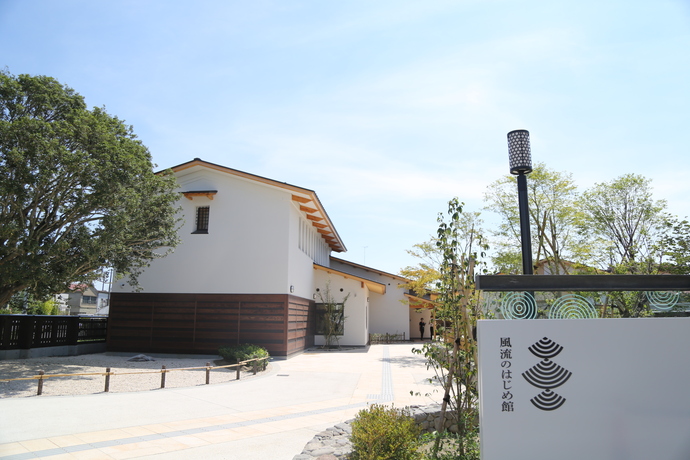 手前に「風流のはじめ館」の名称とロゴが記された白い看板が立ち、その奥に白壁と木材を組み合わせた和モダンな平屋風の建物が広場や樹木と共に佇んでいる、落ち着いた雰囲気の風流のはじめ館の外観写真