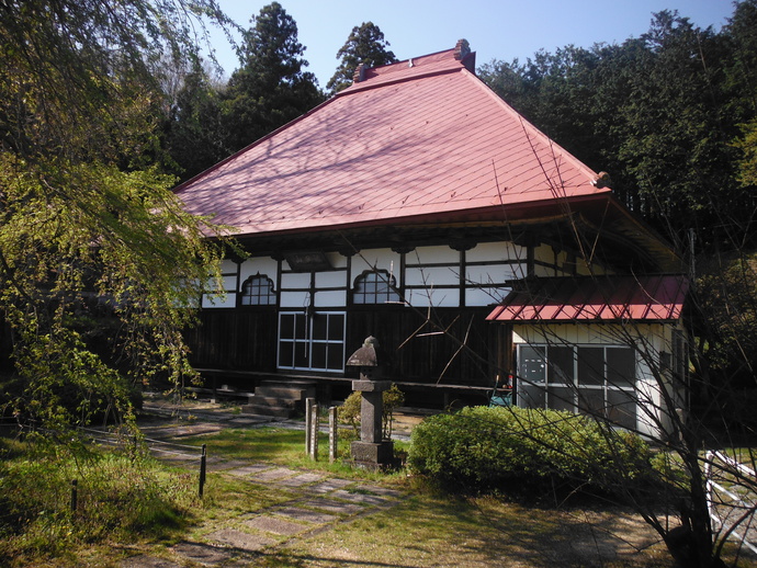 白い壁に黒い柱が映え、赤色の大きな屋根の木造の本堂が緑に囲まれて建ち、手前には石灯籠や手入れされた植栽、石畳の参道が配された護真寺本堂の写真