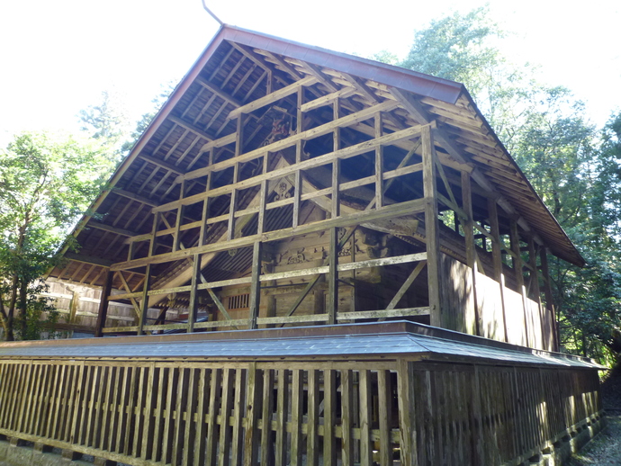 森に囲まれた敷地内に建つ、緻密な彫刻が施された歴史ある桙衝神社の本殿を保護するように、巨大な三角形の屋根を持つ堅牢な木造の覆屋が全体をすっぽりと覆い、周囲には板塀が巡らされている様子を斜め前から捉えた写真