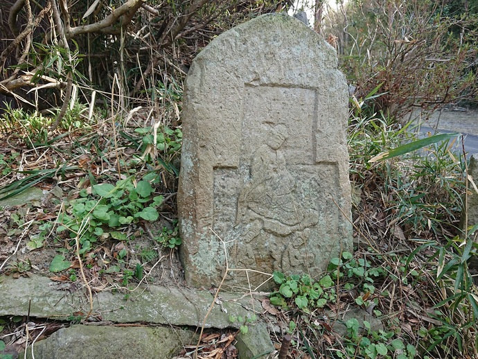 草が生い茂る道端に、烏帽子を被り装束を纏った人物が座っている姿が浮き彫りにされた、古い石碑がひっそりと佇んでいる写真