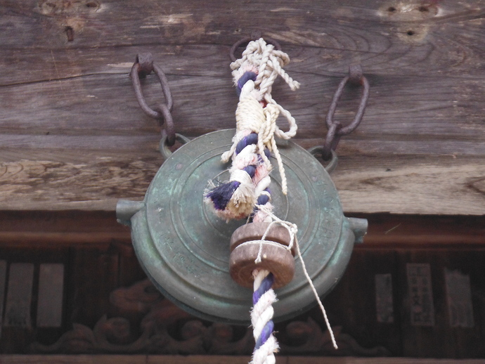 木造建築の梁から錆びた鉄鎖で吊るされた、円盤状で青緑色の錆が浮いた古い鰐口と、その中央に垂れ下がる白と紫の編み込み紐、そして紐に通された木製の撞木を正面から捉えた写真