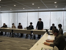 会議室で、L字型に配置した長机の席に複数の男女が着席し、中央に立つ男性が、資料を手にした参加者たちに向かって発言している写真