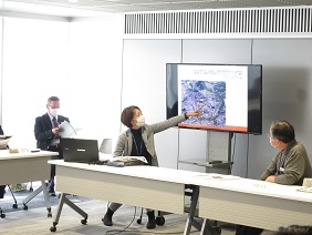 会議室で、マスクを着用した女性がモニターに映し出された空撮画像を指さしながら、参加者に説明をしている写真