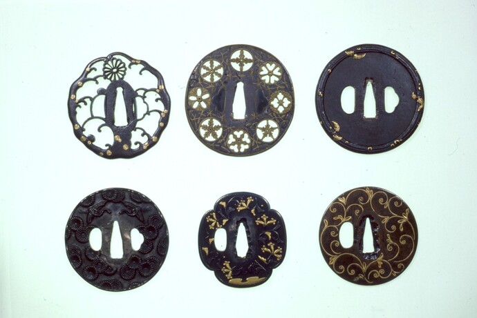 白い背景の上に上下二段に分かれて白や金色の精緻な装飾が施された六つの異なるデザインの鍔が、整然と並べられている写真