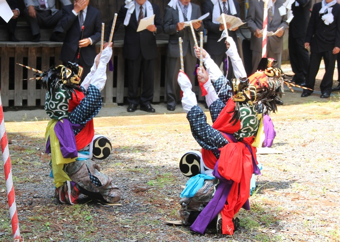 色鮮やかな装束に獅子頭を被った三人の舞い手が、腰に小太鼓を着け、両手に持ったバチを高く掲げながら砂利の上で跪いて舞を披露しており、背後ではスーツ姿の男性たちがその様子を見守っている、伝統的な祭事の光景を捉えた写真