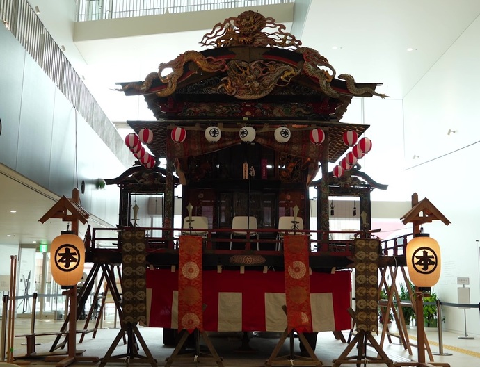 室内の天井が高い展示空間に、提灯や紅白幕が取り付けられた、精緻な木彫と彩色装飾を施した大型の本町屋台が、1対の提灯と共に展示されている様子を正面から捉えた写真