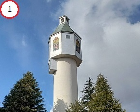 青い空と白い雲を背景に、針葉樹の背後からそびえ立つ白い塔のような形状の建造物と、その上部に設けられたステンドグラス風の窓や小さな鐘楼が特徴的な、晴天下の風景を捉えた写真