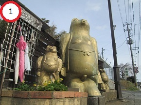 屋外の道沿いに設置された、大小様々なサイズのユーモラスな表情をした狸の置き物が並び、その傍らにはピンク色の傘が立てかけられている写真