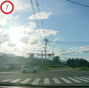 電柱や信号機の向こうに緑豊かな丘が広がる、横断歩道のある交差点を白い乗用車が走行し、白い雲の間から太陽の光が降り注いでいる様子の写真
