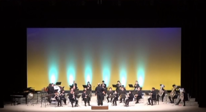ステージ中央で指揮者を前に楽団が円形に配置され、黄から青へのグラデーション照明に包まれて演奏する様子の写真