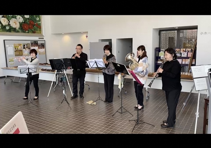明るいロビー空間で管楽器の奏者たちが譜面台を前に横一列に並び演奏を披露している写真