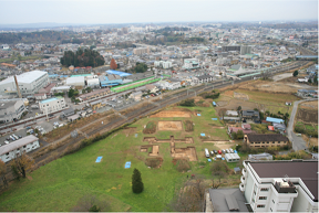 奥にたくさんの建物が建つ町が広がり、手前の広い草地に数箇所土がむき出しになった上人壇廃寺跡が見える写真