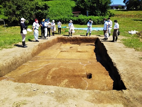 上人壇廃寺跡発掘現場で、掘り下げられた長方形の調査区画の周りに作業服やマスクを着用した複数の人々が立っている見学の様子の写真