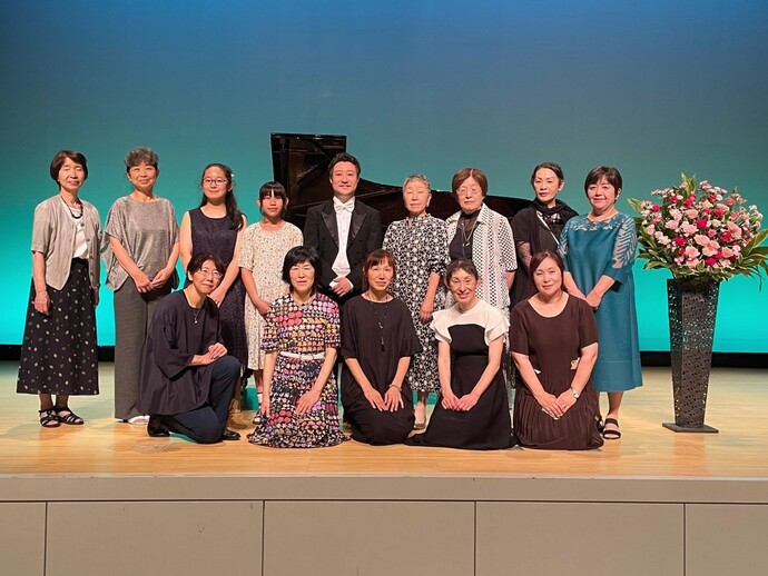 グランドピアノと豪華な生け花が飾られたステージ上に、タキシード姿の男性1人と様々な衣装の女性13人が記念撮影をしている集合写真