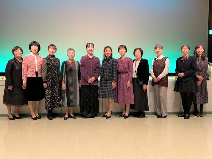 ステージの前で、ワンピースなどの服装をした11人の女性が笑顔で一列に並んで写っている集合写真