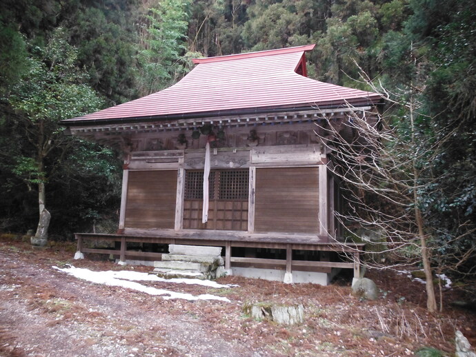 山間の静かな場所に建つ木造の小さな社殿が正面から写り、切妻屋根と格子戸、鈴緒を備えた簡素な造りから地域信仰の場であることが伝わる写真