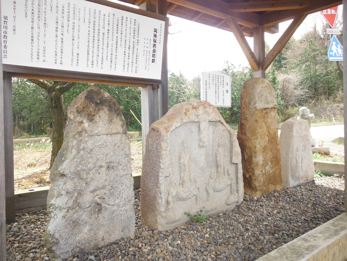木造の屋根の下に、仏像が浮き彫りにされた石碑や自然石を用いた「筑後塚供養塔群」が砂利の上に並び、背後には須賀川市教育委員会による詳細な解説板が設置されている写真