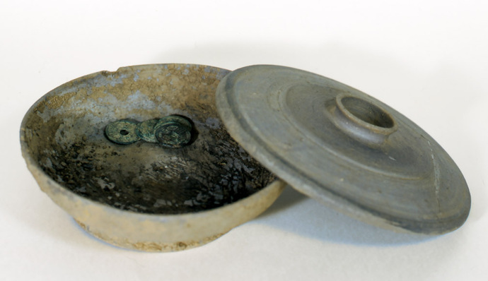 灰色の素焼きのような平皿と蓋が並び、皿の底には経年変化で青緑色に錆びた数枚の古い硬貨が重なるように置かれている、発掘調査によって出土した貴重な遺物を斜め上から捉えた写真