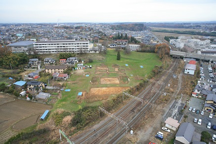 発掘調査が行われている、土がむき出しになった区画が複数ある線路脇の広い草地と、周囲の住宅街や白い大規模な建物を上空から撮影した写真