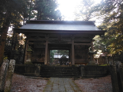 木々に囲まれて石段の上に建つ重厚な木造の随神門で、格子戸を備えた左右の空間と中央の通り道があり、門の奥には本殿が見える様子の写真