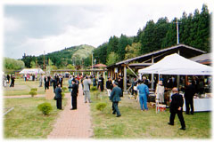 芝生が広がる広場の屋根付きバーベキュー施設の横に白いテントが設置され、その周りに大勢の人々が集まりイベントを楽しんでいる様子の写真