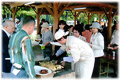 屋根付きのバーベキュー場に大勢の人が集まり、多くの人が集まる屋外の屋根付き休憩所内で、鉄板の上で調理された料理を、白い服の男性が人々の皿に取り分けている様子の写真