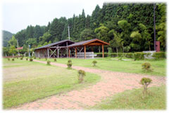 緑の芝生が広がる敷地に、茶色のレンガ敷きの小道がカーブを描きながら続き、奥には森を背景に木造の屋根付きのバーベキュー施設が建っている写真